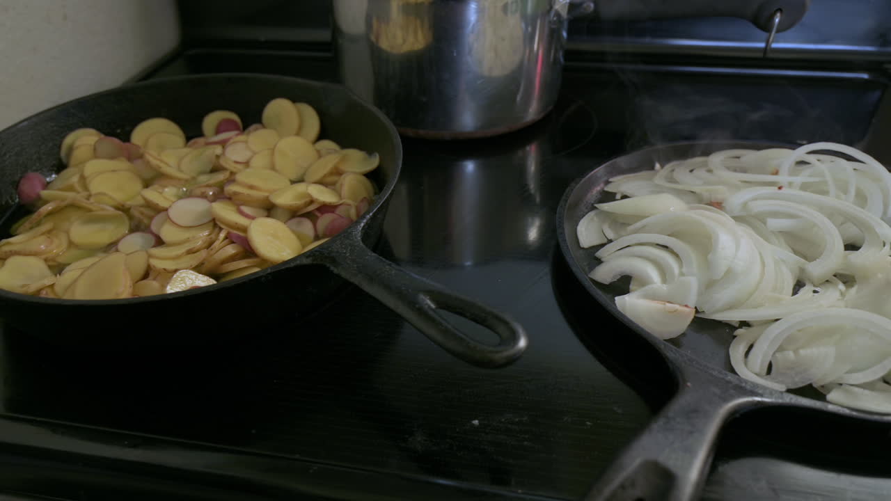 Sliced potatoes and onions cook in two separate frying pans on stove in home kitchen on induction stove