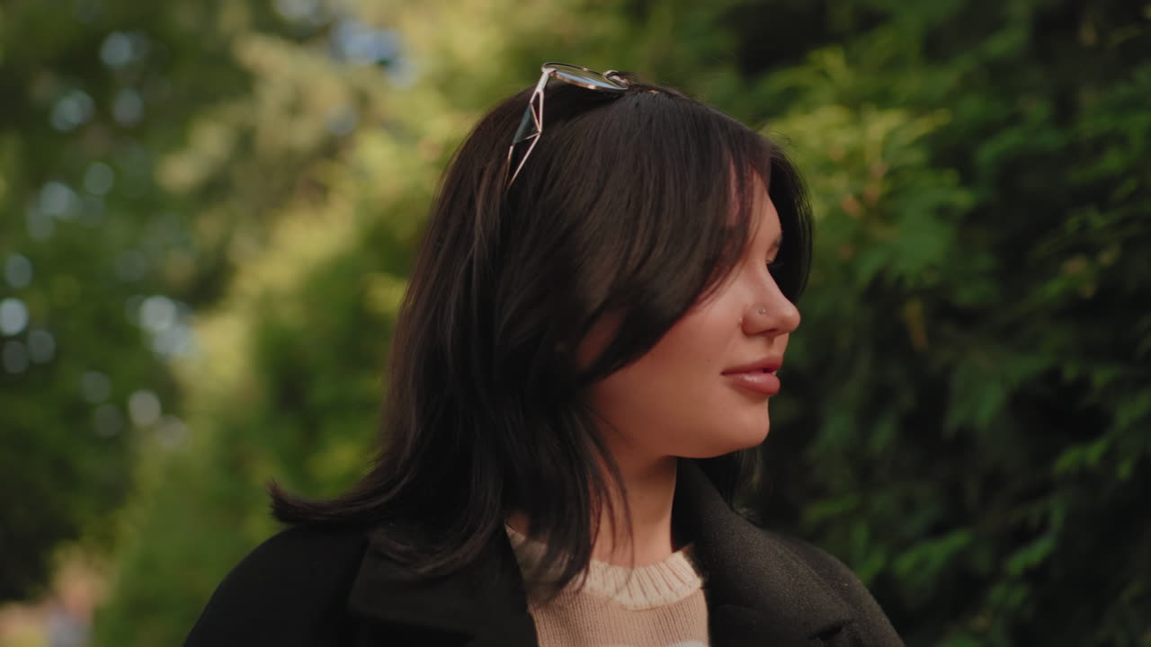 긴 코트를 입은 우아한 여성의 중간 뷰, 녹색 울타리 옆 가을 나뭇잎 위로 손을 뻗어 나뭇잎을 감상하고, 얼굴에는 햇빛이 비치고, 평화로운 산책, 부드러운 보케 배경, 고요한 야외 순간