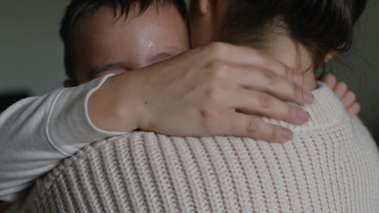 家で、クリーム色のセーターを着た女性が、慰めを求めて顔を押し付け、涙を流す子供を抱きしめ、背中をさすっている。