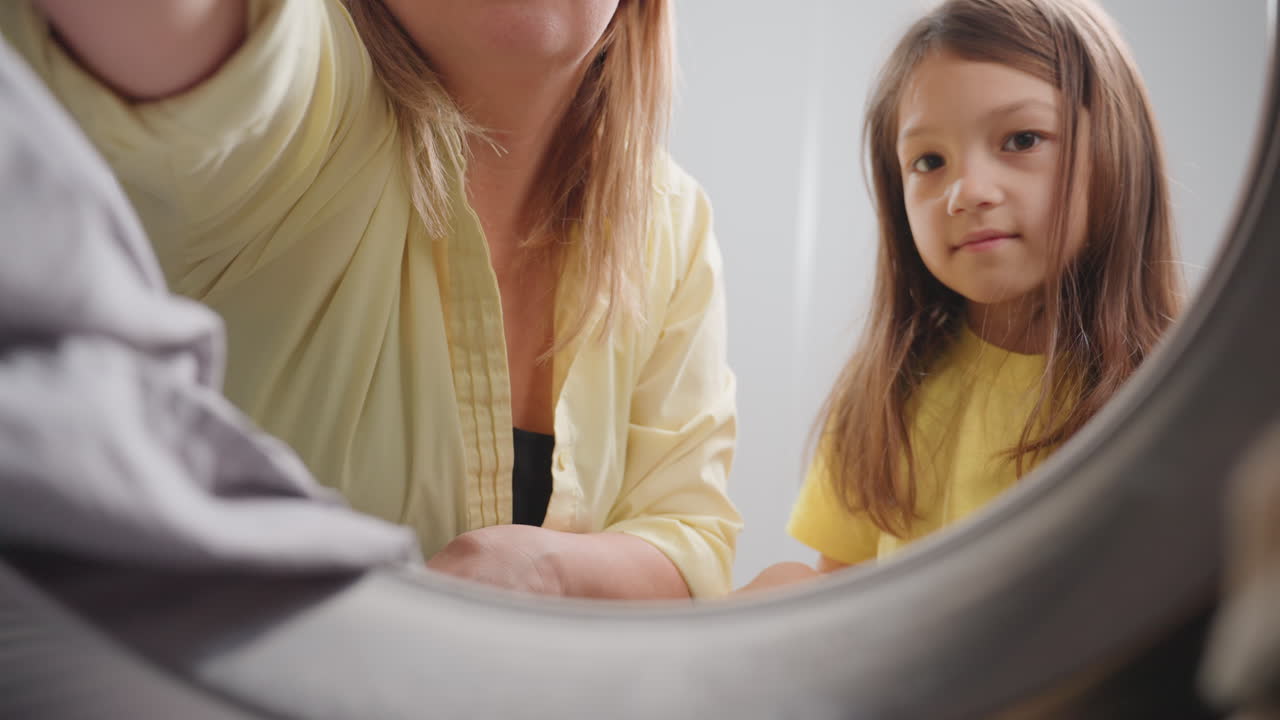 Inside washer pov shows mother with toddler loading fabrics through door, careful hands reach toward lens, teamwork during laundry chore, family hygiene moment, soft light, modern home