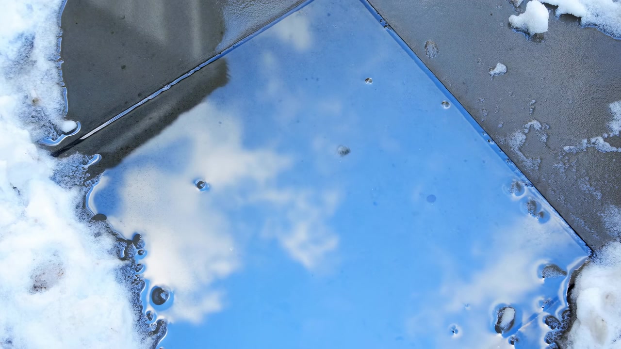 Early sequence frame evolving toward motion, with a reflective puddle centered in the frame and bordered by snow on dark wet asphalt