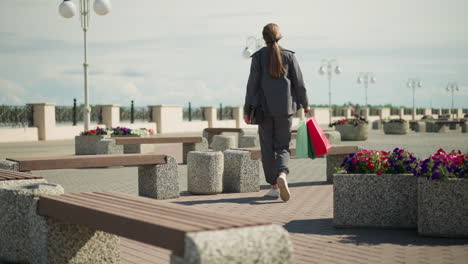 lady holding three shopping bags walks towards outdoor benches, places the bags on the bench, and adjusts her black handbag while seated, the background includes flowers and benches