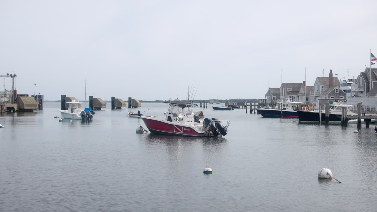 Håndholdt opptak av båter som ligger i Nantucket havn, Massachusetts, med brygger og sjønære hus under et mykt, overskyet kystlys.