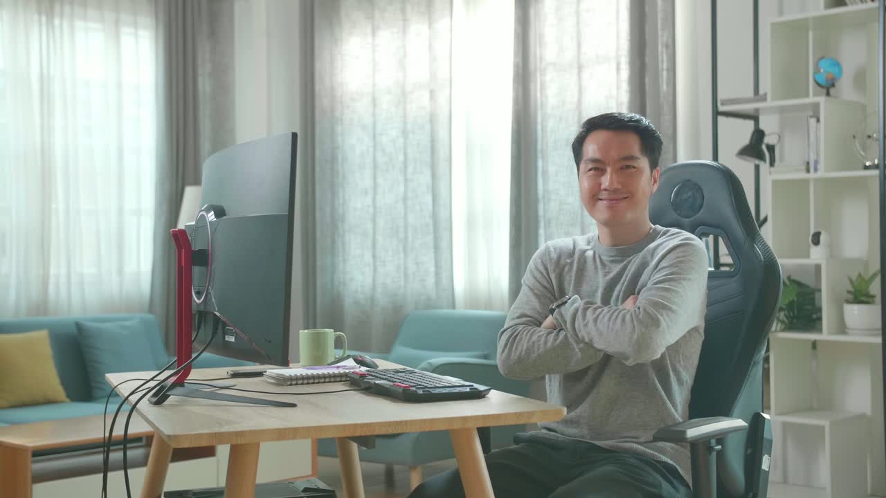 Asian Man In Long Sleeved T-Shirt And Black Pants Crossing His Arms And Smiling To Camera While Working With Desktop Computer At Home.