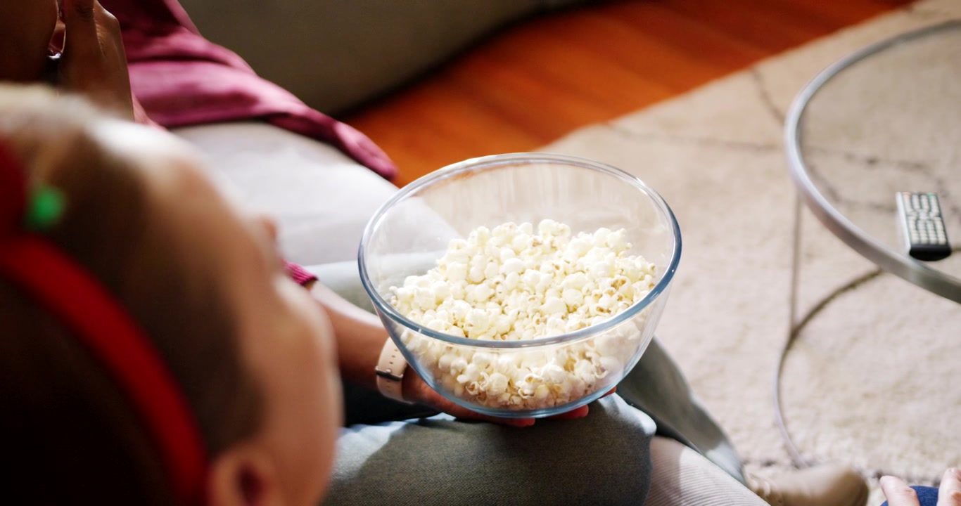 Bambino seduto sul divano con cuffie rosse, ciotola di popcorn in grembo che fa merenda mentre la TV è accesa.