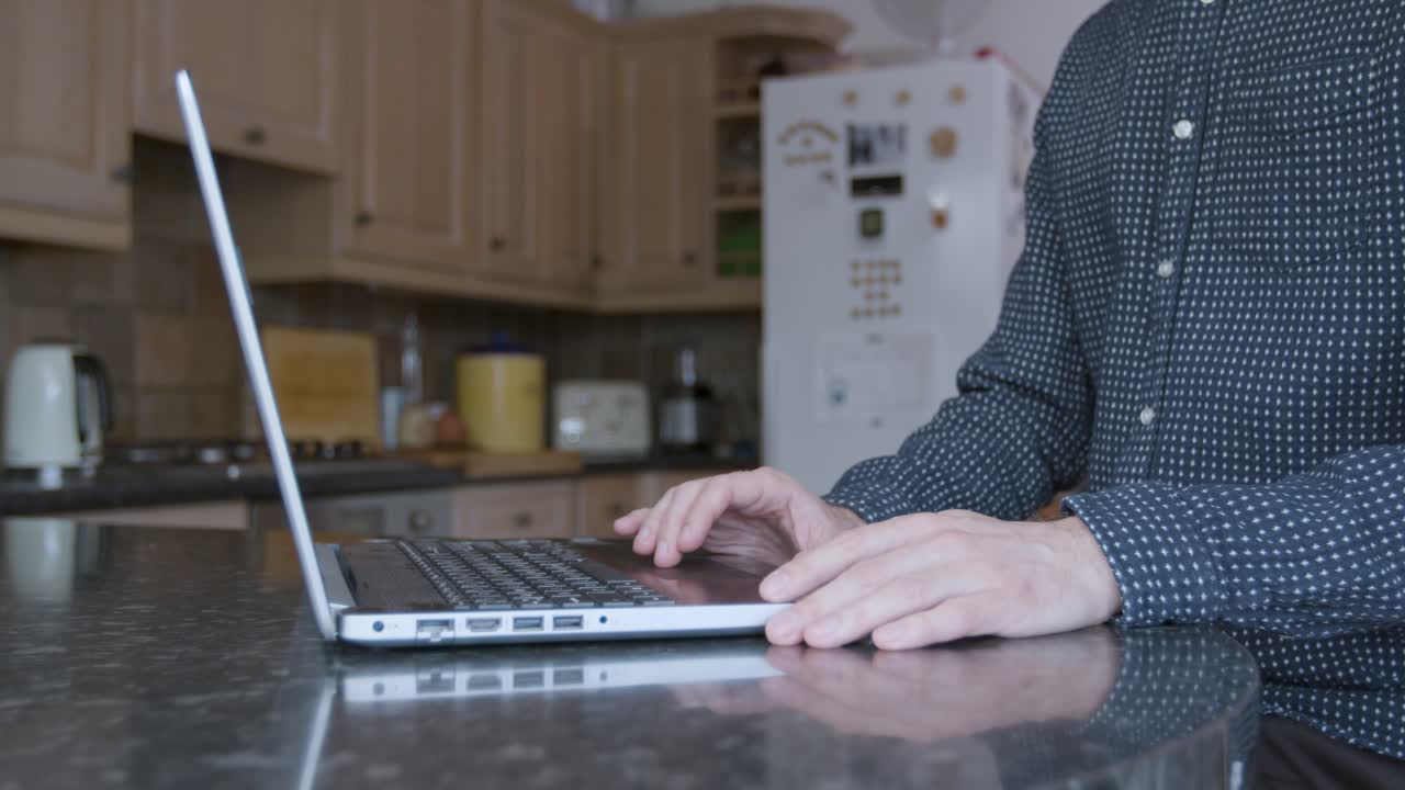 Smartly dressed man using the track pad and typing on a laptop on a kitchen counter while working from home