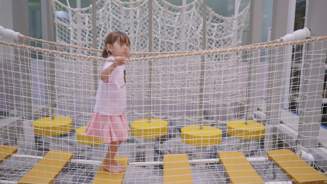 Toddler girl walking and hopping along bouncing bridge in indoor play area, showing balance and coordination as she jumps to each platform, medium sideview static