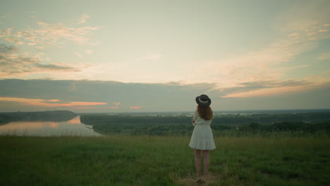 a woman in a white dress and hat stands thoughtfully in a grassy field by a tranquil lake at sunset. she gazes into the distance, embracing the calm and reflective moment in nature's serene setting