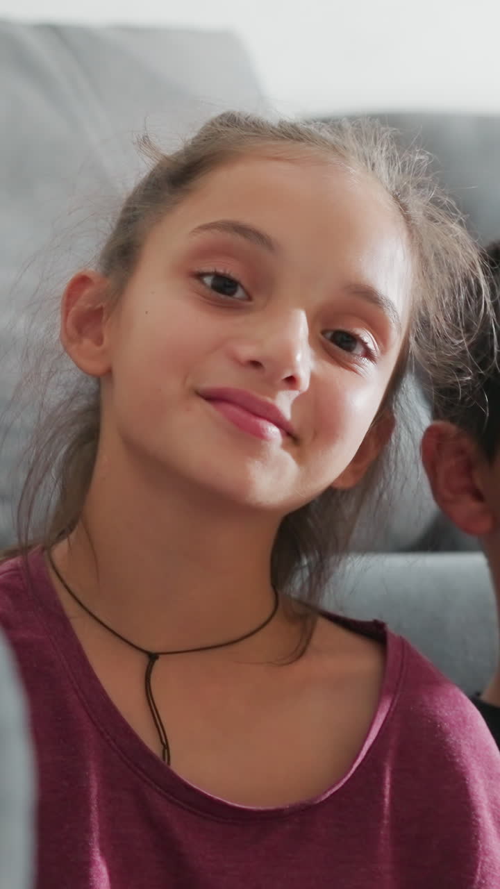 Una niña adorable apoya suavemente la cabeza en el hombro de su hermano mientras ambos se sientan juntos en el suelo del salón, apoyados en un sofá mullido, mirando a la cámara con expresiones sutiles que capturan el tierno vínculo fraternal.