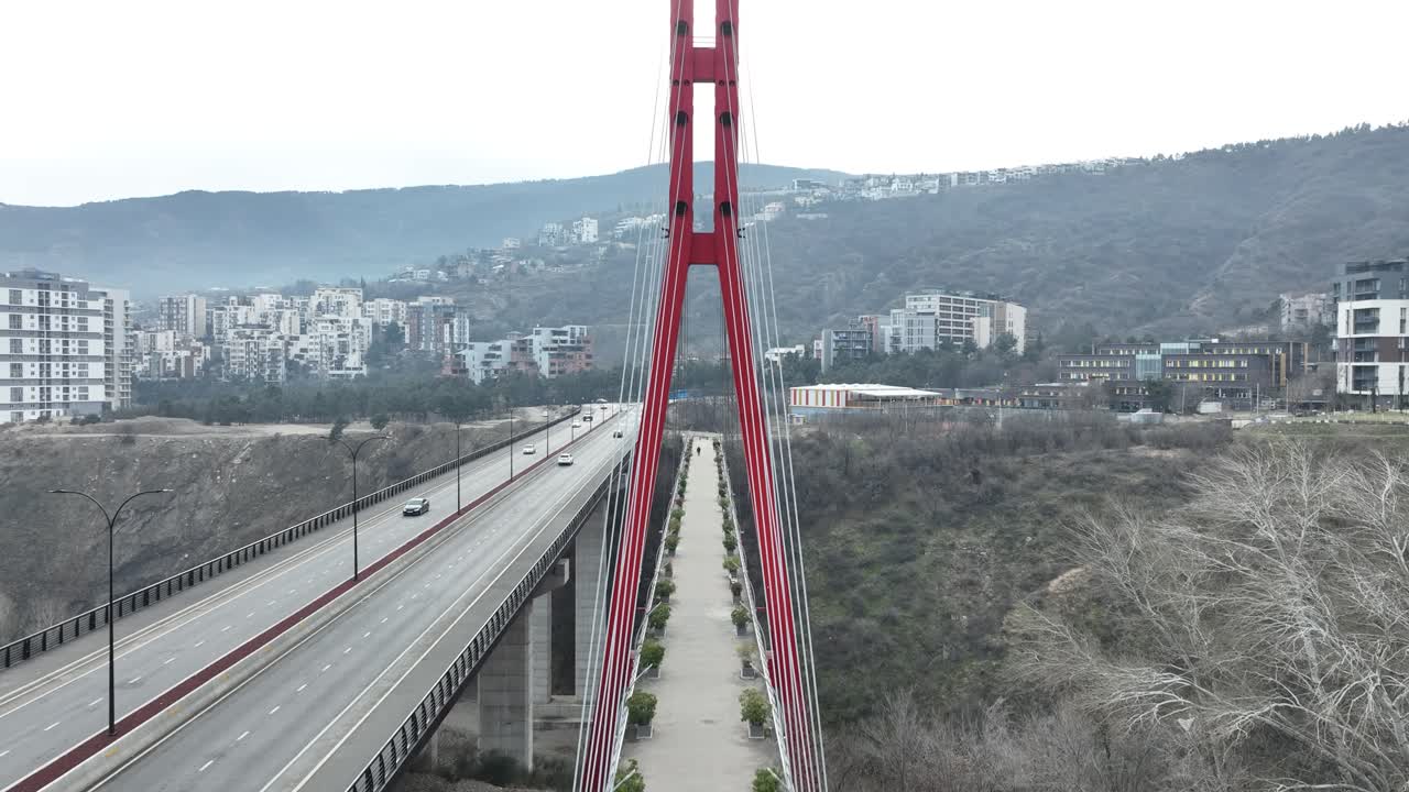 A striking red suspension bridge spans a green valley near a bustling urban area. The structure stands out against the surrounding cityscape, blending nature with urban infrastructure