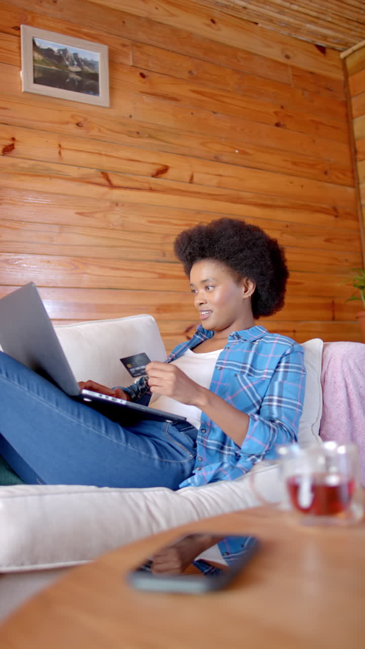 Vertical video of african american woman using laptop and credit card in living room, slow motion
