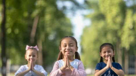 three young girls performing the traditional thai greeting wai, pressing their palms together with a slight bow, smiling in a park setting, demonstrating respect and cultural heritage