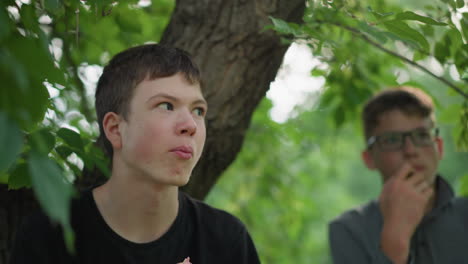 close-up of young enjoying a snack under a tree, he appears focused on something in the distance while another person in the background is blurred, also eating under a leafy tree