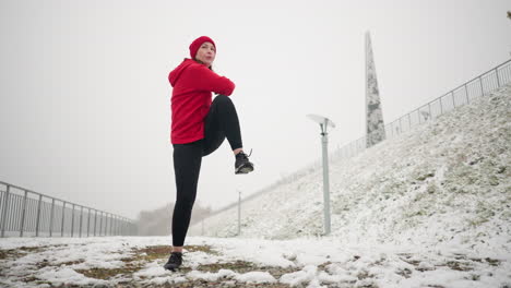 mujer realizando un enérgico entrenamiento de invierno al aire libre, levantando su pierna en el suelo nevado mientras usa una sudadera roja con capucha y guantes negros, en contra de una ladera nevada y un fondo de niebla sereno