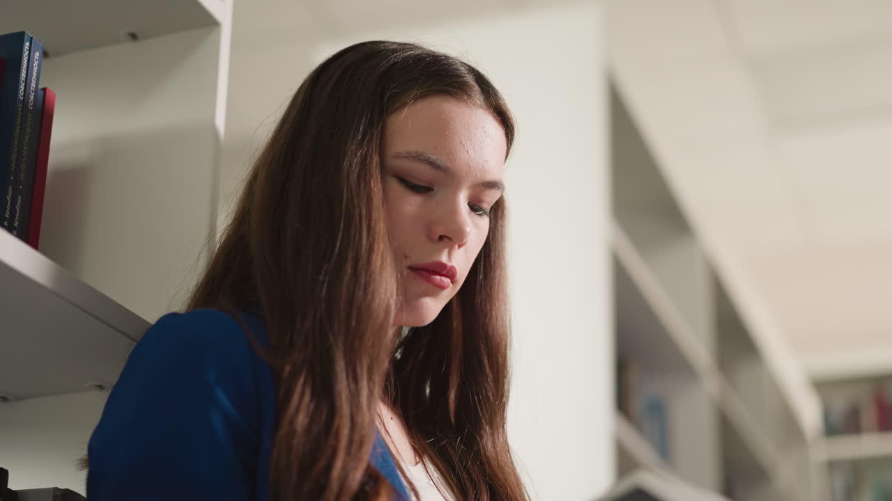 mujer concentrada se apoya en la estantería de libros de primer plano. dama morena mira hacia abajo de pie en la librería de primer plan. estudiante visita el almacenamiento de literatura para materiales