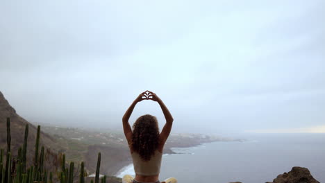 en la cima de una montaña, una mujer medita y emplea el gesto de la mano maha sakal, con el océano y las montañas verdes como telón de fondo