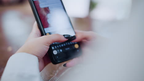 close-up of black cell phone being used by woman to chat, hands typing on screen, placed on wooden table with vibrant blurred light effect in the background