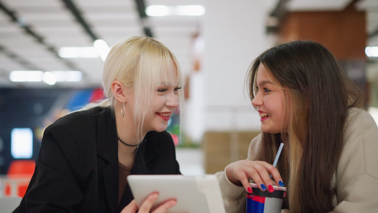Makeup artist proudly presents her latest makeup design on tablet to friend at cafe table while friend reacts with surprise and excitement, holding drink, sharing beauty creativity