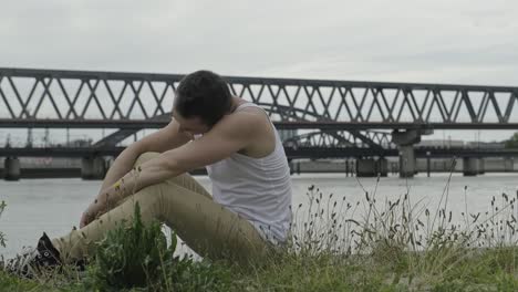 side view of positive young male in casual outfit rotating head and looking at camera with smile while sitting against modern urban bridge crossing river