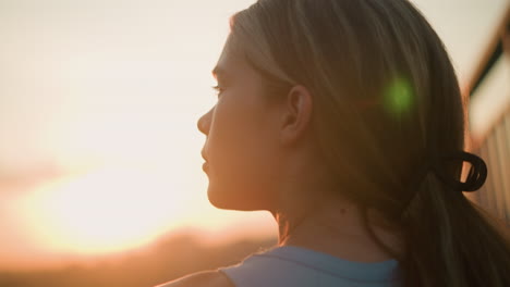 close-up of young lady with thoughtful expression, sunlight reflecting off her face, sitting near iron railing at dusk while gazing into distance