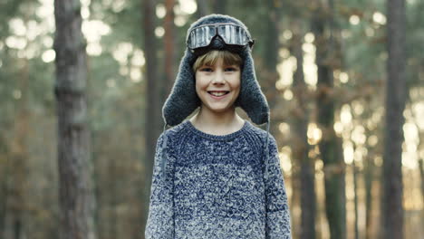 pequeño niño caucásico soñador de ser piloto con sombrero y gafas especiales sonriendo a la cámara en el bosque