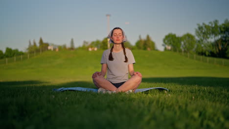 a smiling young woman sits on her yoga mat, her glowing skin lit by golden evening sun. a green hill rises behind her and a soft shadow dissolves on the grass as the camera moves smoothly to the left.