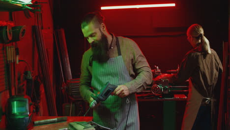 caucasian young man welder with long beard and apron working with electrodrill and drilling in workshop