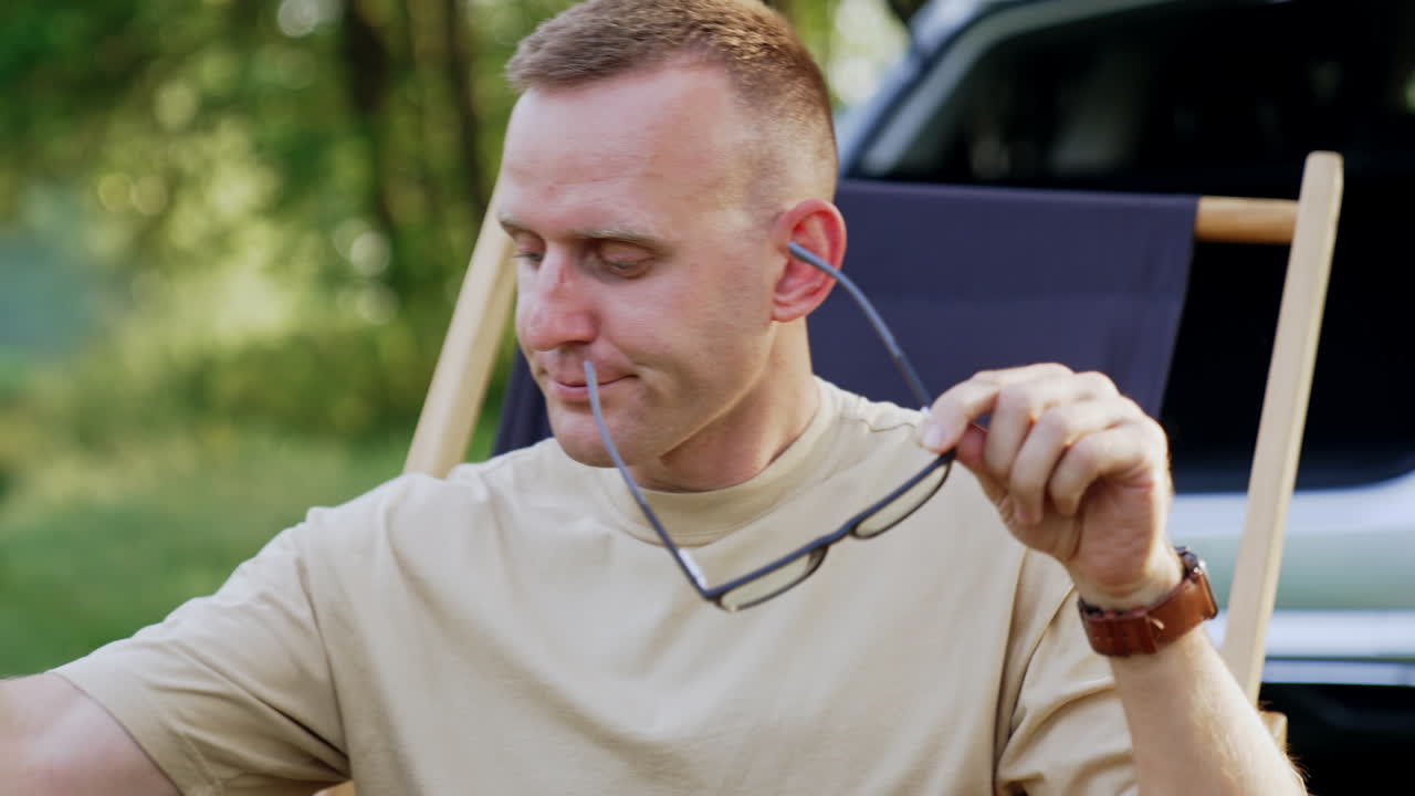 Tired mid-aged man finishes work on laptop. Freelancer puts off his glasses and sits back in folding chair relaxed. Beautiful summer forest at backdrop.