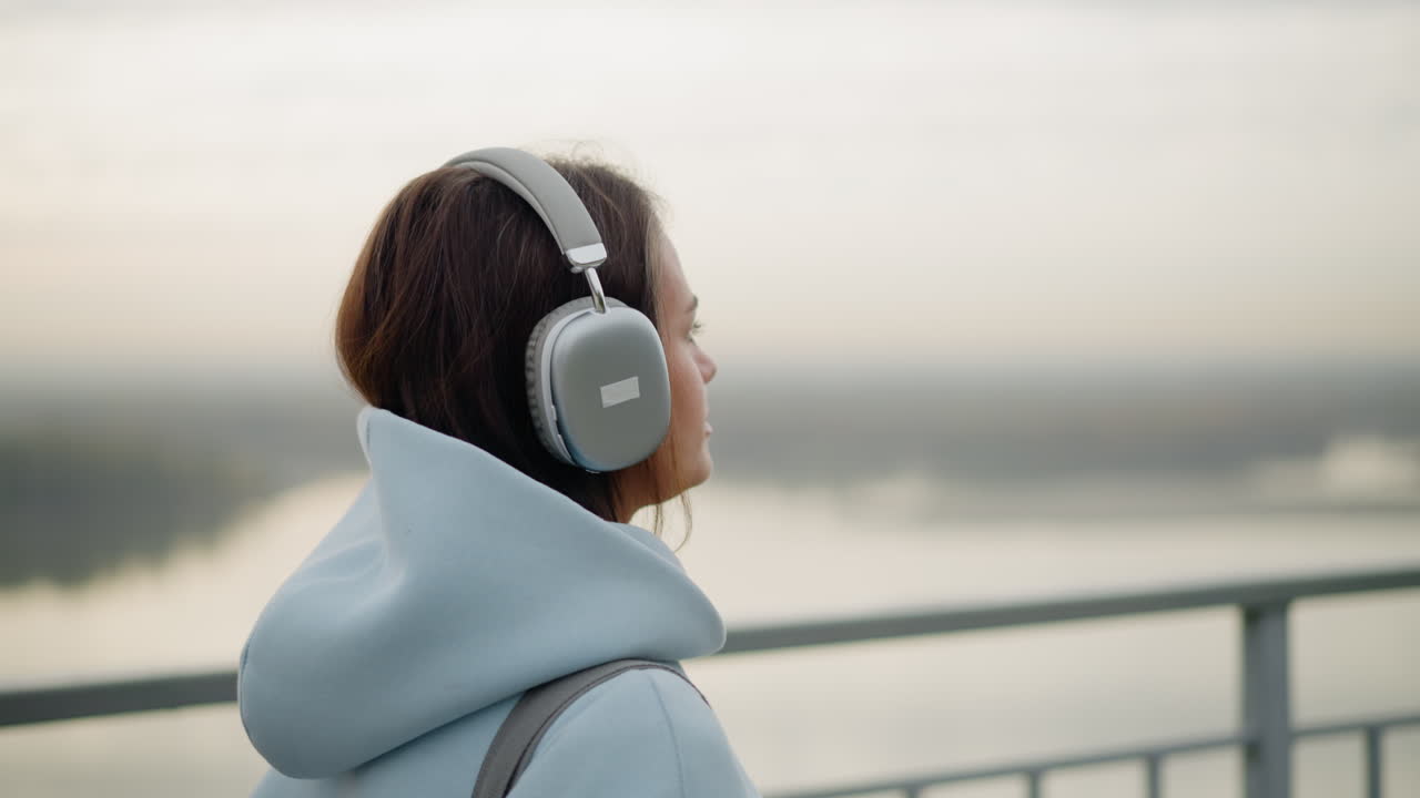 Close-up side view of young female student listening to music with headphones as she strolls near iron rail beside flowing water in peaceful outdoor setting, calm, relaxing walk by river