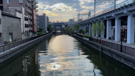 Sunset-reflects-on-a-calm-canal-in-a-city,-flanked-by-buildings-and-greenery