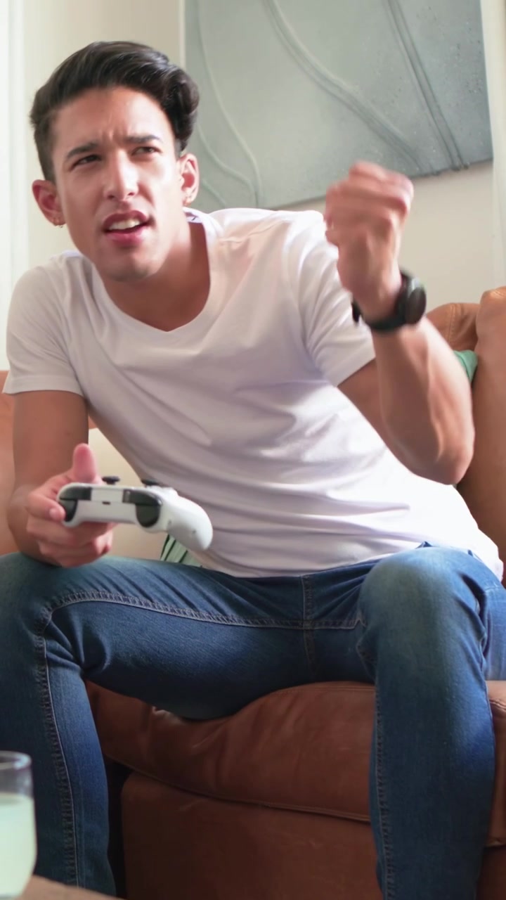 Video verticale: Sporgendosi in avanti, uomo che tiene un gamepad bianco sul divano, fa uno spuntino e reagisce al gioco.