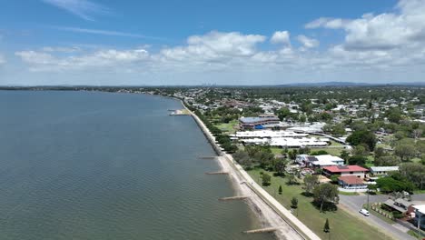 Drone-establishing-pull-away-shot-of-Brisbane-Brighton-Suburb,-camera-flying-over-water-Moreton-Bay-with-Brisbane-City-in-background