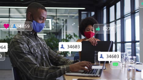 Animation-Von-Social-Media-Symbolen-über-Einem-Mann-Mit-Gemischter-Abstammung,-Der-Eine-Gesichtsmaske-Trägt-Und-Im-Büro-Einen-Laptop-Benutzt