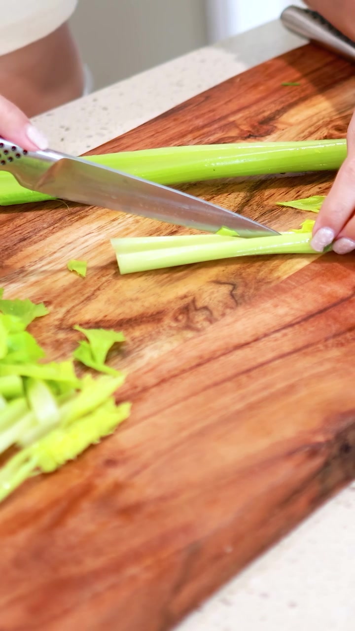 Mani che affettano il sedano su un tagliere di legno in una cucina luminosa. Le inquadrature ravvicinate evidenziano la tecnica di taglio precisa e gli ingredienti freschi.