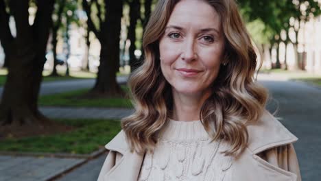 mature woman radiating serenity while standing in sunlit park, wearing elegant beige coat and white sweater, expressing genuine happiness through warm, confident smile