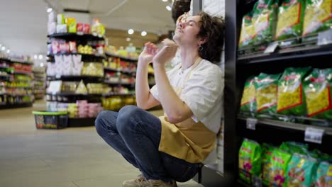 Un-Chico-Cansado-Con-El-Pelo-Rizado,-Trabajador-De-Un-Supermercado,-Se-Apoya-En-La-Pared-Cerca-De-Los-Estantes-Con-Productos-Y-Se-Toma-Un-Descanso-Durante-Un-Día-Ajetreado-En-El-Trabajo.