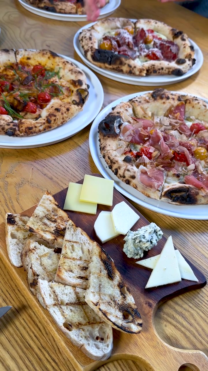 Vista dall'alto con zoom indietro di un tagliere di formaggi e pane grigliato accanto a pizze italiane gourmet fresche