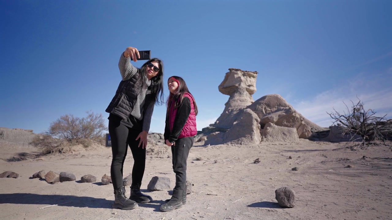 Due ragazze posano per un selfie contro le formazioni rocciose uniche del Parco Provinciale di Ischigualasto, Argentina. Piena luce del giorno, abbigliamento casual, grandangolo, cielo azzurro limpido, atmosfera di turismo avventuroso