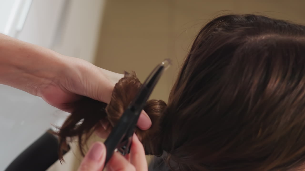 헤어 스타일리스트가 머리를 스타일링하고 있고, 백인 여성이 머리 스타일링을 받고 있으며, 젖은 머리를 묶어 고정하고 있습니다. 스타일리스트는 샴푸대 근처에서 트리트먼트를 위해 젖은 머리를 정돈된 묶음으로 조심스럽게 고정하고 있습니다.