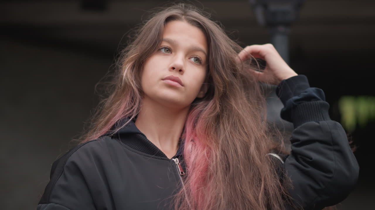 Teen adjusting hair under bridge aspiring dancer frames face with wet pink strands, black hoodie, confident gaze, casual streetwear attitude, subtle raindrops and soft overcast light, motion captured