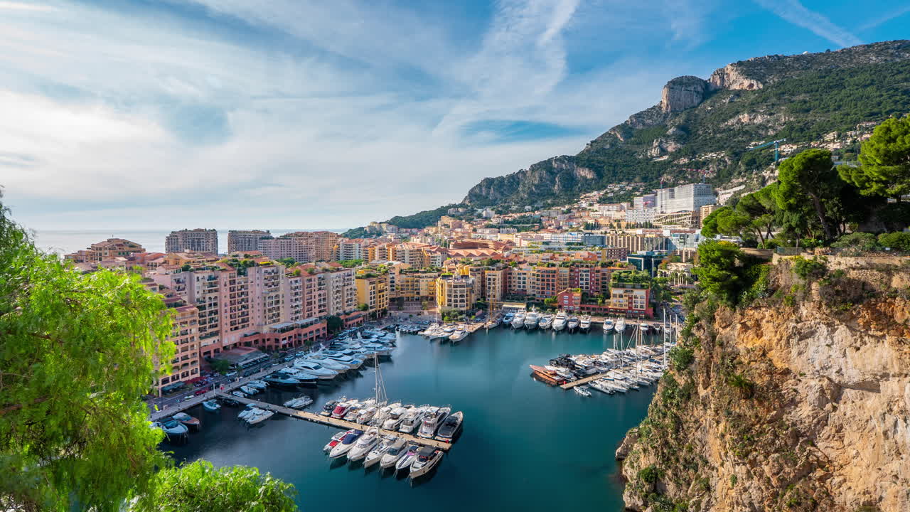 Timelapse diurno del pittoresco Porto di Fontvieille a Monaco, con yacht di lusso ormeggiati nel porto turistico circondato da edifici colorati e scogliere rocciose.