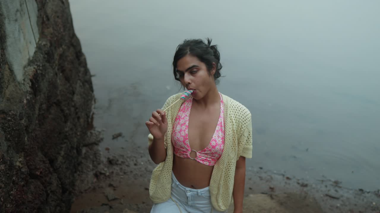 Uma mulher indiana está sentada no mar saboreando um picolé. Ela está usando um top de biquíni rosa e um suéter branco.