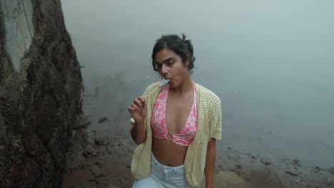 A-sexy-indian-woman-woman-is-sitting-on-the-sea-and-licking-lollypop-ice-cream-She-is-wearing-a-pink-bikini-top-and-a-white-sweater