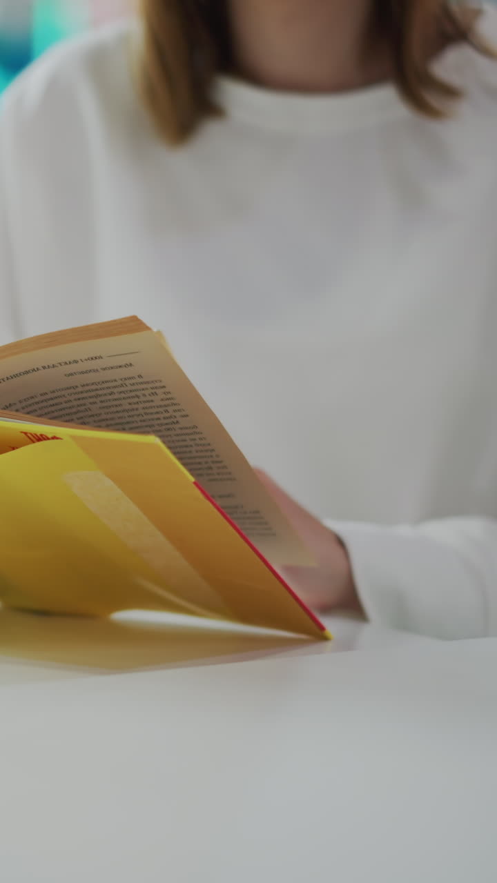 primer plano de manos hojeando las páginas de un libro amarillo en una mesa con una taza de café cerca, iluminación suave y fondo borroso con juguetes coloridos y decoración vibrante en un entorno relajado de un centro comercial