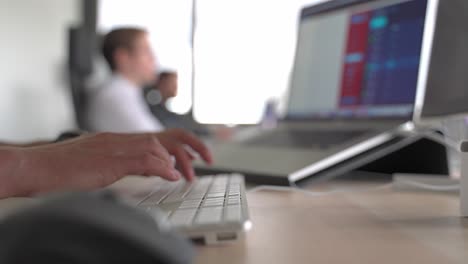 Office-sitaution-male-hand-typing-on-a-silver-keyboard-with-notebook-screen-and-collegues-in-the-background