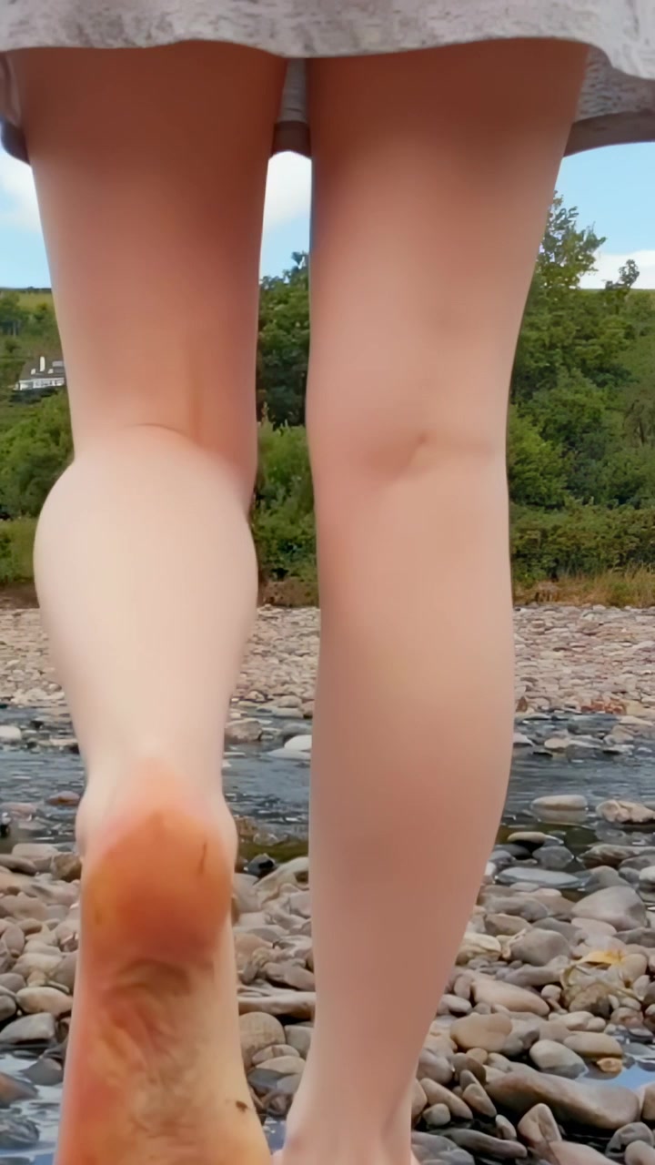 Video verticale: Donna che cammina sollevando il piede, a piedi nudi sui sassi verso un ruscello, con una gonna corta.