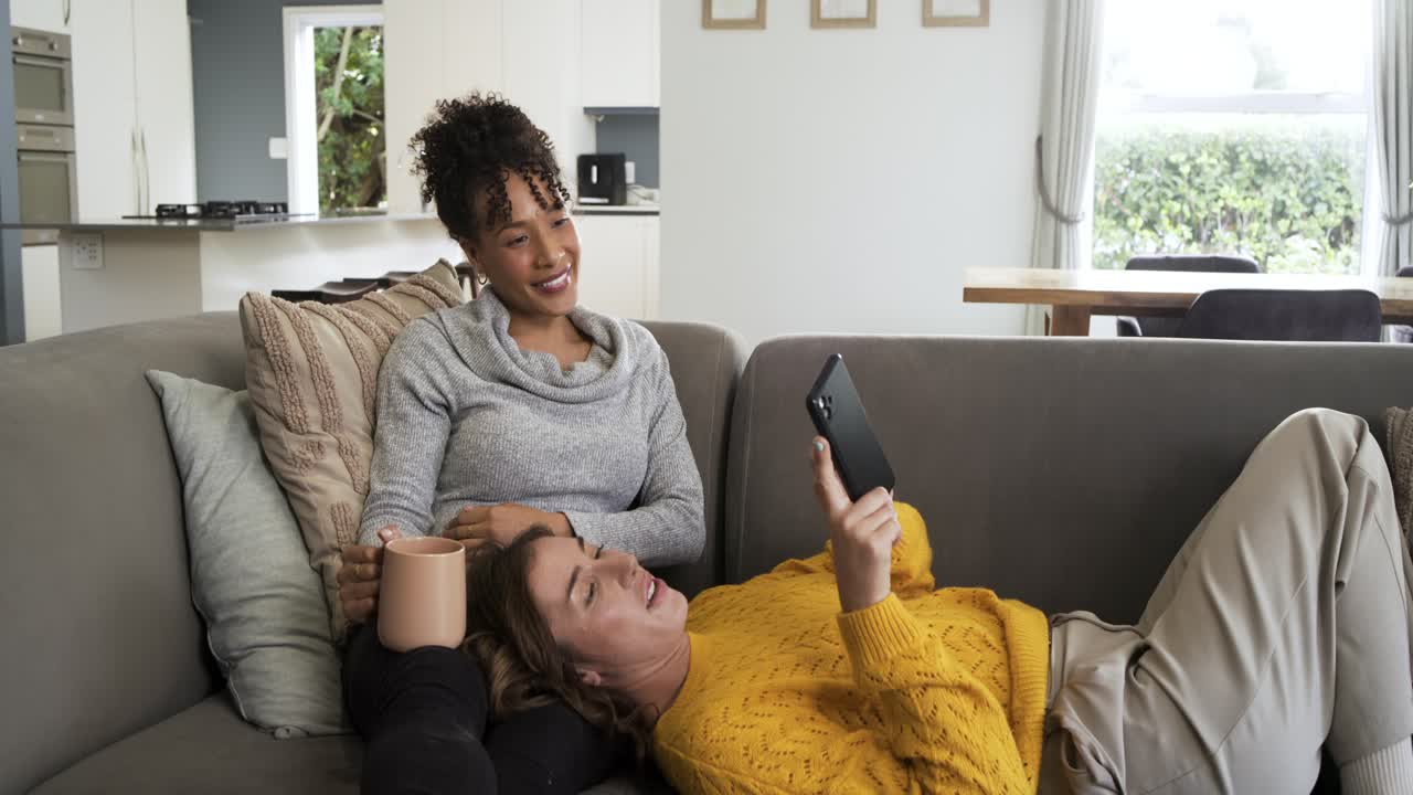 After tablet lift two women lying on living room sofa swiping tablet and sipping coffee
