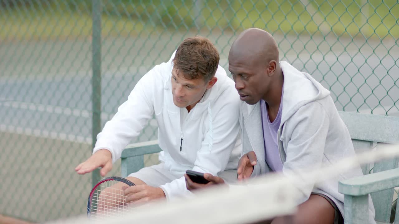 Felici amici uomini di diverse origini che guardano lo smartphone seduti su una panchina al campo da tennis, al rallentatore.