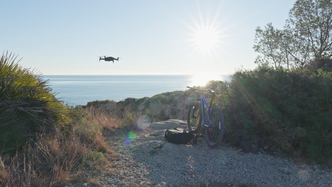 Drone entering rocky coastal scene where mountain bike is parked with morning sun rising over blue ocean