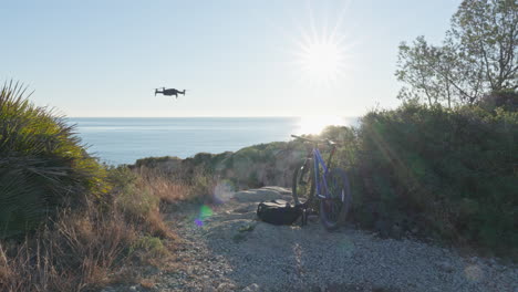 drone entering rocky coastal scene where mountain bike is parked with morning sun rising over blue ocean